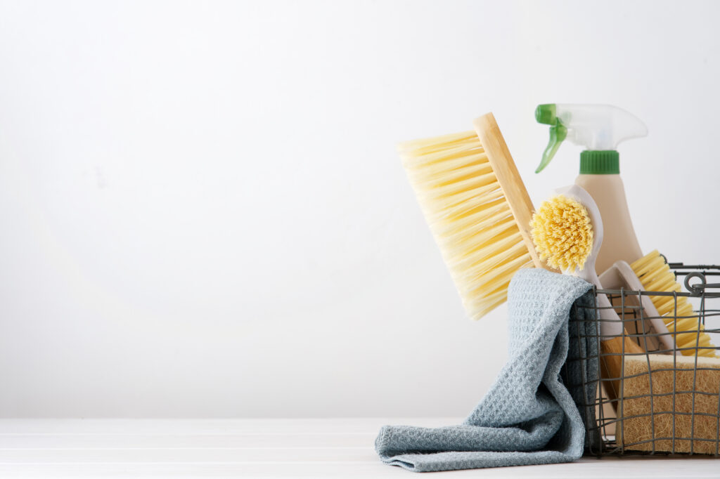 Assortiment de produits de nettoyage sur une surface blanche, comprenant un balai, des brosses, une bouteille de nettoyant à vaporiser, et un chiffon, tous rangés dans un panier en métal.