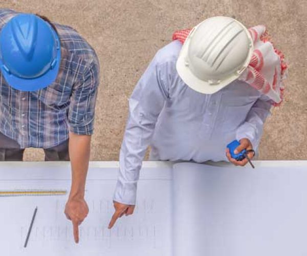 Deux assistants à maîtrise d'ouvrages, portant des casques de sécurité, analysent des plans de construction sur une table.