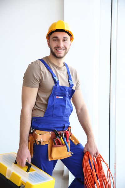 Young smiling electrician holding bunch of wires and tool box indoors