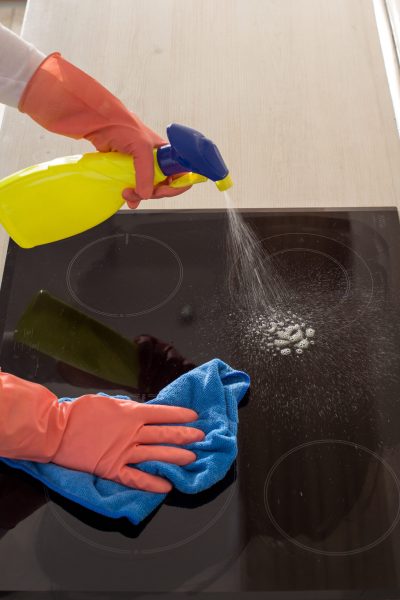 Close up of female hands with protective gloves spraying induction stove and cleaning with wipe in kitchen