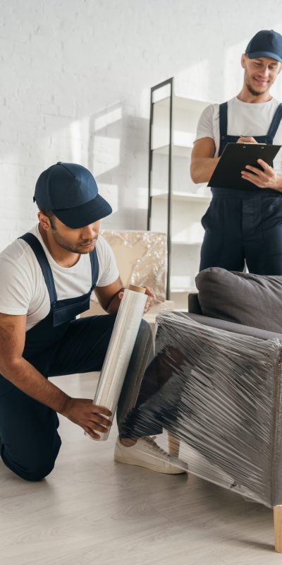 Indian mover in cap holding stretch film roll near sofa and worker with clipboard on blurred background