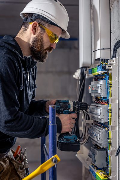 A male electrician works in a switchboard with an electrical connecting cable, connects the equipment with tools.